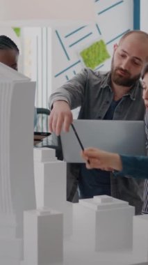 Dikey video: Tablet ve dizüstü bilgisayarla masadaki bina modelini analiz etmek için çalışan çok ırklı insanlar. Mimarlık projesi için inşaat yapısı ve yerleşimi tasarlamak için teknolojiyi kullanan meslektaşlar.