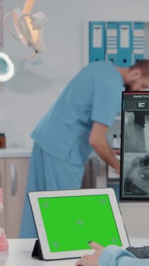 Dikey video: Oral bakım ve diş sağlığı için tablet üzerinde yatay yeşil ekran kullanan ortodontist. Krom anahtarı olan dişçi olarak çalışan bir kadın ve diş taramasına bakarken izole edilmiş bir model şablonu.