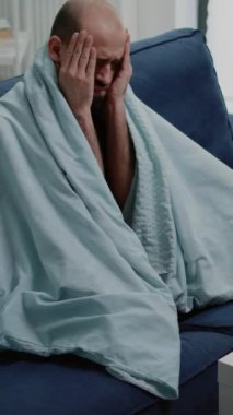 Dikey video: Baş ağrısı olan, şakakları ovalayan ve battaniyeye sarılmış hasta hisseden hasta bir adam. Mevsimsel koronavirüs salgını nedeniyle grip ve grip belirtileri gösteren hasta bir yetişkin. Hastalıklı kişi