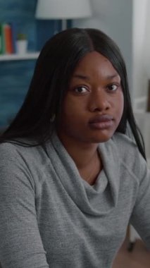 Dikey video: Oturma odasında masada oturan Afro-Amerikan öğrencinin portresi ev soğutma sırasında gülümseyen kameraya bakıyor. Siyahi gençler lise bilgilerini tarıyor.