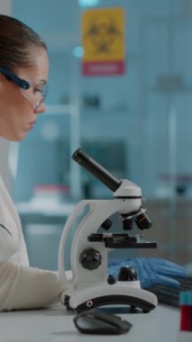 Dikey video: Laboratuvarda DNA örneğini incelemek için mikroskop kullanan bilim mühendisi. Laboratuvar aletleri ve bilgisayarında mikroskobik cam lenslerle çalışan koruyucu gözlük takan bir kadın biyolog.