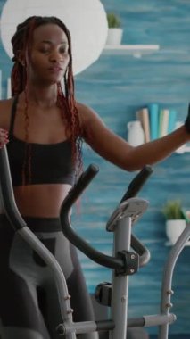 Verical video: Spor salonunda spor yaparken eliptik bisiklete binip koşan zinde siyah kadın oturma odasında sağlık rutin fitness videosu izliyor. Atletik siyah kadın vücut kaslarını çalıştırıyor.