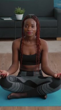 Verical video: Siyah kadın portresi lotus pozisyonunda oturma odasında sabah nefes egzersizi yapıyor. Sporcu kıyafeti giyen zayıf, formalı yetişkin vücut kaslarını esnetiyor.