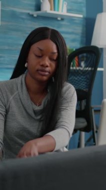 Dikey video: Afro-Amerikalı öğrenci blogcusu kulaklıkları evden uzakta, sosyal medya enginar tarama bilgi konferansında bilgisayar başında çalıştırıyor. Fasulye çuvalında oturan Afrikalı kadın.