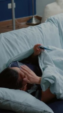 Dikey video: Kanepede yatan hastalıklı kadın ve termometre kullanarak ateşi ölçen kadın. Hasta yetişkin, masada mendil ve ilaç şişeleri varken cihaza bakıyor ve sıcaklığı kontrol ediyor.