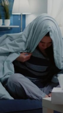 Dikey video: Evde sıcaklık ölçümü için termometreyi kullanan kişi battaniyeye sarılmış. Üşüyen ve titreyen bir kadın ateşini kontrol ediyor. Yetişkinlerin titreme ve virüs belirtileri var..