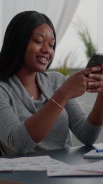 Dikey video: Evinden uzakta masa başında otururken modern telefon kullanarak siyah tenli bir öğrenci mesaj gönderiyor. Sosyal medyaya bakan Afro-Amerikalı bir kadın yaşam tarzı tavsiyelerini paylaşıyor.