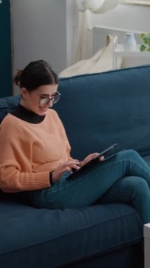 Dikey video: Uzak okul işleri için dijital tablet kullanan bir kız öğrenci, kanepede oturuyor. Üniversite araştırması yapan ve proje ödevi hakkında notlar alan bir kadın çevrimiçi kurs için rapor hazırlıyor.