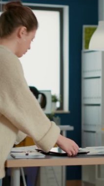 Woman holding digital tablet zooming in on sales pie chart and comparing data with statistics on laptop screen and papers on desk. Startup employee working with key performance indicators in office.