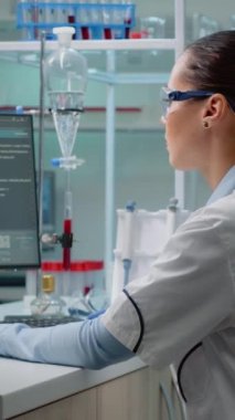 Dikey video: Laboratuvarda hücre animasyonu ile modern bilgisayar kullanırken petri kabını sıvı bakterilerle inceleyen yenilikçi bilim adamı. Laboratuvar önlüklü ve gözlüklü kadın eczacılık yapıyor.