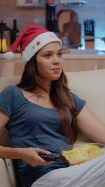 Dikey video: Noel Baba şapkası takan ve Noel arifesinde atıştırmalıklarla televizyon izleyen neşeli yetişkin. Dekorasyon alanında elinde uzaktan kumandayla televizyona bakıp kanal değiştiren neşeli bir kadın.