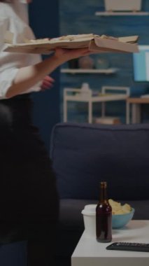 Dikey video: Oturma odasında televizyon izlerken birisi akşam yemeği için masaya bir kutu pizza koyuyor. İşten sonra yemek yemeye hazırlanan bir kadın evde kanepede yalnız oturuyor.