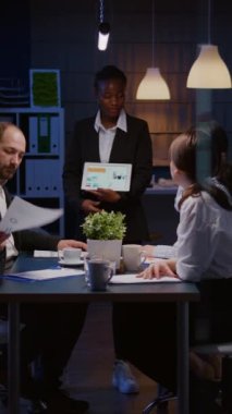 Dikey video: Tükenmiş işkolik Afrikalı Amerikalı şirket toplantı odasında aşırı çalışan tablet kullanarak finansal grafikler gösteriyor. Çok ırklı iş adamları yönetim istatistiklerini tartışıyor