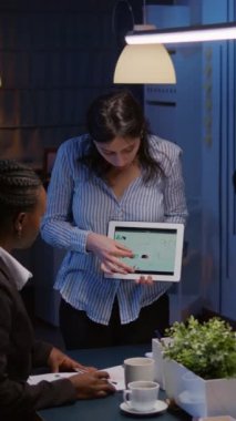 Dikey video: Çok çalışan işkolik girişimci kadın, gece geç saatlerde şirket çözümünde çok çalışan tablet kullanarak pazarlama grafiklerini şekillendiriyor. Çeşitli etnik çeşitlilikte takım çalışması çözümü