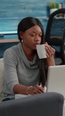 Dikey video: Koyu tenli blogger öğrencisi oturma odasında kahve içerken evde iletişim bilgilerini tarıyor. Medyada evden çalışan Afrikalı kadın
