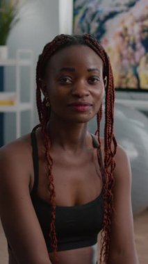 Verical video: Yoga haritasında Lotus pozisyonunda oturmuş oturma odasındaki fitness antrenmanında çalışan, zinde siyah bir kadının kameraya bakan portresi. Afrikalı zayıf yetişkin vücut kasları ısınıyor