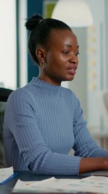 Portrait of african american woman typing using laptop keyboard and smiling at camera sitting at desk in busy startup office. Casual business employee working relaxed with sales statistics.