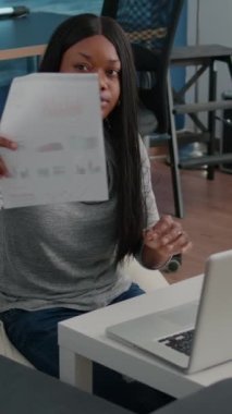 Dikey video: Evden uzakta çalışan Afro-Amerikan öğrenci bilgisayardaki sunum stratejisini analiz eden finansal grafikler yazan pazarlama istatistikleri. Siyah kadın fasulye çuvalında oturuyor.