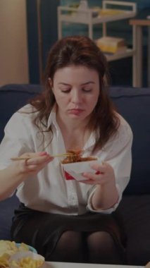 Dikey video: Oturma odasında oturan kişi yemek çubuklarıyla Asya eriştesi yiyor. Çin yemeği paketinden beyaz kadın TV ekranında film izlerken yemeğin tadını çıkarıyor.