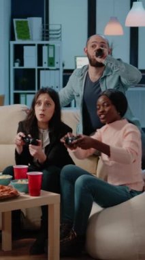 Dikey video: Neşeli iş arkadaşları TV 'de joystickler ve konsollarla oyun oynuyorlar. İş arkadaşları TV 'deki denetçilerle oyun kazanıyor, işten sonra içki ve atıştırmalıkların keyfini çıkarıyorlar.
