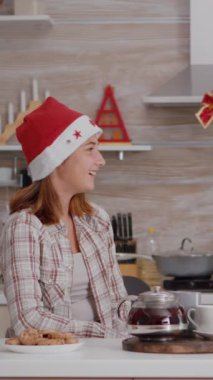 Dikey video: Büyükannenin geleneksel kış tatilinde hediye paketi ile torununa sürpriz yapması süslü xmas mutfağında eğlenmesi. Noel Baba şapkalı mutlu aile kutlaması.