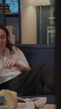 Dikey video: Masada hazır yemek yerken kanepenin üzerinde cipslerle rahatlayan beyaz yetişkin videosu. Kanepede oturup TV 'de film izleyen bir kadın.