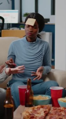 Dikey video: Afrikalı Amerikalı kadın işten sonra yapışkan notlarla oyun tahmin etmeye çalışıyor. Meslektaşlar, ofiste bira ve pizzayla kutlama yaparken eğlenceli aktiviteler yapmak için tahmin oyunu oynuyorlar..