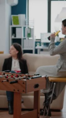 Dikey video: Erkek, kadınla langırt masasında futbol oynuyor, işten sonra içki ve eğlence için iş yerinde buluşuyor. Çalışma saatleri dışında bira ve pizzanın tadını çıkaran çok ırklı bir grup arkadaş.