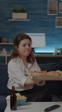 Dikey video: Oturma odasındaki masada pizzacı adamdan bir kutu pizza getiren kişi. Fast food ve alkollü içecekle kanepede oturup televizyon izleyen genç bir kadın. Yetişkin siparişi.