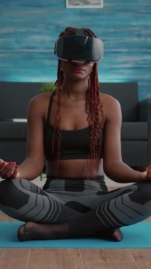 Verical video: Oturma odasında meditasyon yaparken yoga haritasında Lotus pozisyonunda otururken sanal gerçeklik kulaklığı takan zinde siyah bir kadın. Sağlıklı yaşam tarzının tadını çıkaran bir yetişkin.