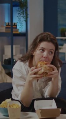Dikey video: Beyaz kadın TV 'de kanepede oturmuş hamburger ve gazoz yudumluyor. Paket yemek yiyip meyve suyu içen ve televizyon izleyen biri. Yetişkinler sağlıksız bir akşam yemeği yiyor.
