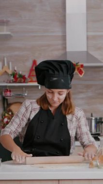dikey video: Önlük takan mutlu bir çocuğun portresi ev yapımı hamuru rulo broşla yapıyor geleneksel lezzetli zencefilli tatlı hazırlıyor. Xmas dekore edilmiş mutfakta masada duran kız.