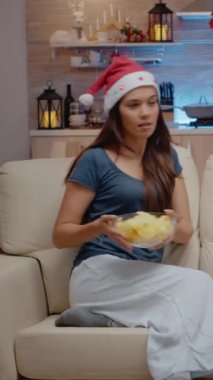 Dikey video: Şenlikli kadın televizyon izliyor ve Noel süslemeli mekanda kaseden cips yiyor. Yılbaşı tatilinde atıştırmalık yerken uzaktan kumandayı kullanan kişi kanal değiştiriyor