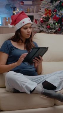 Dikey video: Tablet üzerinde çalışan ve Noel arifesinde televizyona bakan şenlikli kişi. Noel Baba şapkası takan bir kadın kış tatilini kutlamak için dijital cihaz kullanıyor ve televizyonda film izliyor.