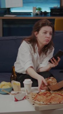 Dikey video: Pizza yiyen ve koltukta oturan akıllı telefon ekranına bakan kişi. İnternet teknolojisi olan beyaz bir kadın oturma odasında hazır yemek yerken televizyon izliyor.