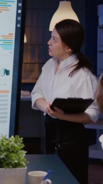 Dikey video: İşkolik iş kadını, ofis toplantı odasında fazla mesai yapan sunum monitörünü kullanarak şirket çözümünü sunuyor. Finansal istatistikleri analiz eden çok ırklı takım çalışması