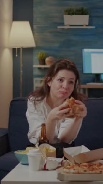 Dikey video: Oturma odasında lezzetli hamburger ve patates yiyen kişi alkollü içecek içip televizyon izliyor. Fast food servisi yapan bir kadın kanepede oturup dinleniyor.