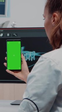 Dikey video: Dişçi ofisteki bilgisayardaki diş taramasına bakarken dikey olarak yeşil ekranlı akıllı telefonu tutuyor. İzole edilmiş olanlar için krom anahtarı olan cep telefonu kullanan diş bakımı uzmanı
