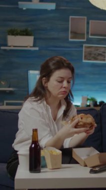 Dikey video: İşten sonra evdeki kanepede oturan genç kişi paket burger ve biradan hoşlanıyor. Oturma odasında televizyon izlerken kadın fast food yiyor ve alkollü içecek içiyor.