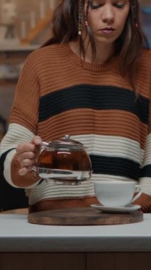 Dikey video: Beyaz kadın Noel 'de mutfakta oturmuş çaydanlıktan çay dolduruyor. Mevsimlik bayram kutlaması için hazırlanırken kış içeceğiyle ısınan genç insan