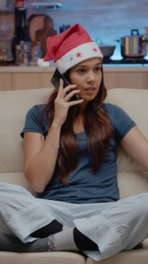 Dikey video: Uzak noel kutlaması için ailesiyle akıllı telefondan konuşan şenlikli kadın. Kırmızı Noel Baba şapkalı yetişkin telefonda sohbet ederek televizyon izliyor ve cips yiyor.