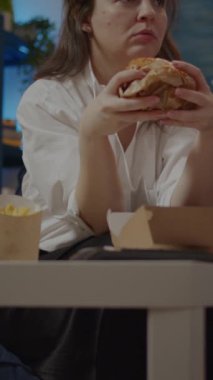 Dikey video: Oturma odasında yemek siparişi veren yetişkin videosu. Kadın evde kanepede otururken hamburger yiyor ve biradan bira içiyor. Teslimatı seven kişi