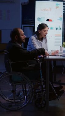 Dikey video: Tekerlekli sandalyedeki çok çalışan engelli işadamı, gece geç saatlerde şirket iş ofisinde çalışırken görmezden geliniyor. Çok ırklı iş adamlarının beyin fırtınası yaptığı işkolikler