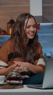 Dikey video: Noel 'de dekore edilmiş odada dizüstü bilgisayarla konuşan şenlikli kadın. Geleneksel bayramı kutlayan genç yetişkinler evde internet üzerinden çay dolduruyorlar.