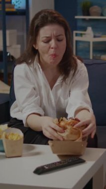 Dikey video: Yetişkinlerin hamburger yiyip patates kızartması yemesi oturma odasında televizyon izlemesi kanepede oturması. Fast food yemeği olan genç bir kadın, paket servis sonrası evde soda yiyip içmenin keyfini çıkarıyor.