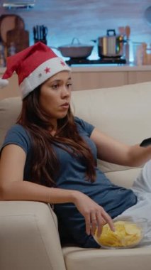 Dikey video: Noel Baba şapkalı bir kadın televizyon kumandası kullanarak kanal değiştiriyor ve televizyon izliyor. Sıkılmış insan Noel arifesinde ekrana bakar ve kaseden cips yer. Yalnız bir yetişkin.