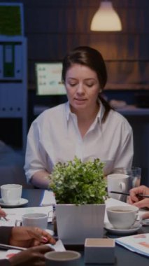 Dikey video: Çeşitli etnik çeşitlilikte takım çalışması ofis toplantı odasında gece geç saatlerde mali grafikleri analiz ediyor. İş arkadaşları proje fikirleri üreterek şirketleri çözüyor.