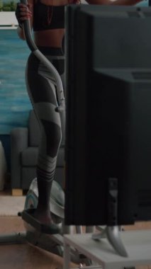 Verical video: Spor giyim bölümünde zayıf formda bir kadın antrenör sabah aerobik antrenman sırasında spor salonunda eliptik bisiklet kullanarak vücut kaslarını çalıştırırken televizyonda fitness videosu izliyor. Atletik sağlığın tadını çıkarıyor