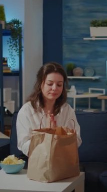Dikey video: Genç bir kadın oturma odasında televizyonu açarken yemek paketini masaya koyuyor. Kafkasyalı kişi işten sonra evde televizyon izlerken teslimat yemeği hazırlıyor