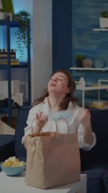 Dikey video: Genç bir kadın dışarıdan yemek aldıktan sonra maskesini çıkarıyor, oturma odasındaki masaya torba koyuyor. Beyaz adam TV izlerken sağlıksız yemek yemeye hazırlanıyor.